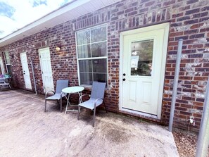 Un bâtiment en briques avec une porte blanche, deux fenêtres et un petit patio comprenant deux chaises et une table.
