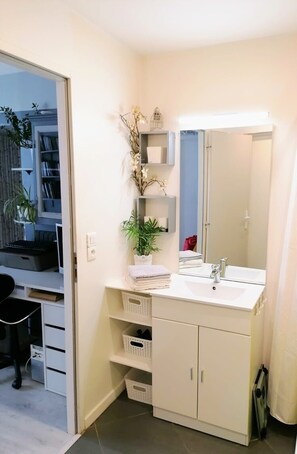 A modern bathroom with a white sink, mirror, and cabinets. There are shelves with decorative items and a potted plant.
