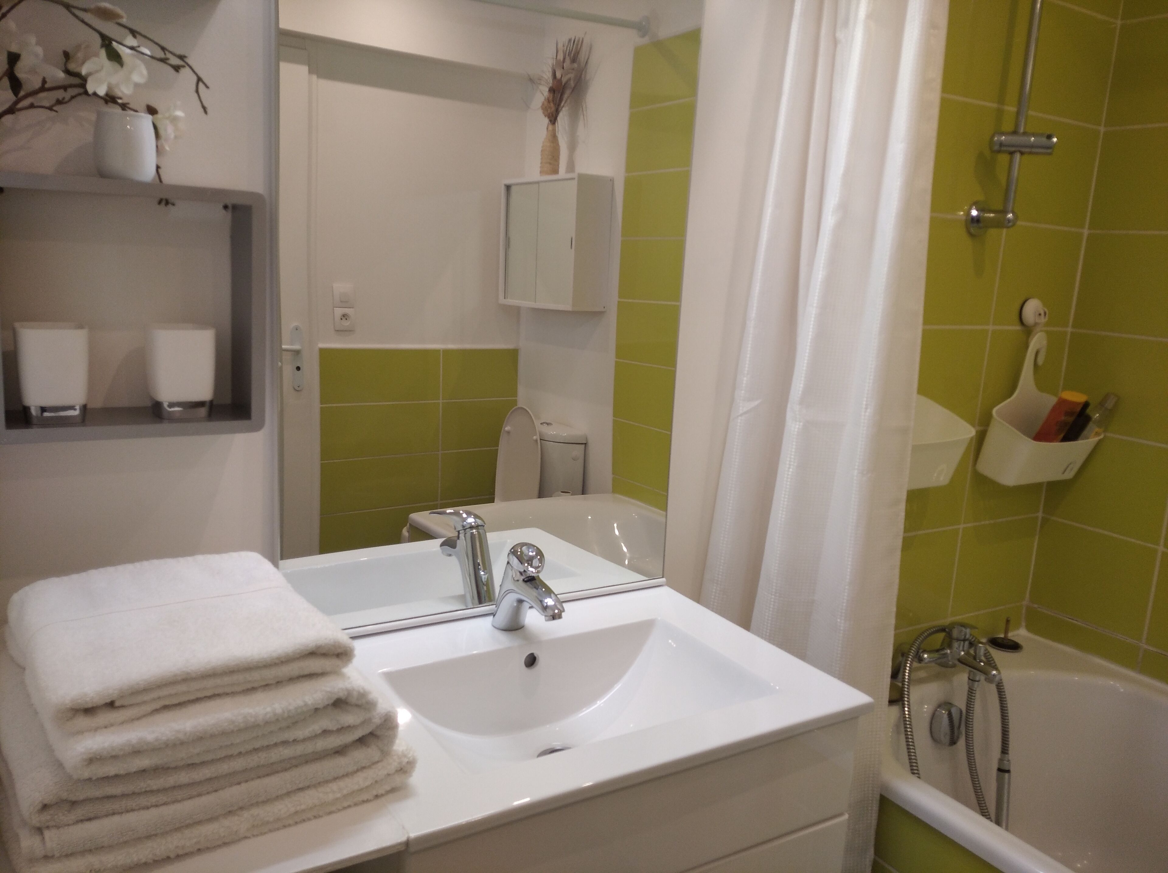 Une salle de bain avec un lavabo blanc, un miroir, une pile de serviettes blanches, une douche avec une porte en verre et des murs recouverts de carreaux verts.