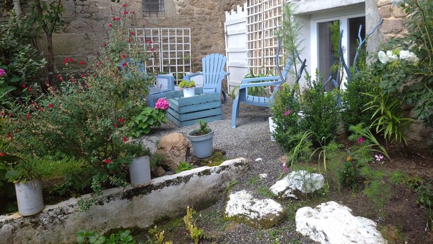 A stone building with a garden area featuring a seating arrangement with blue chairs and a table, surrounded by various plants and flowers. - Appartement Nature et Jardin (Eymoutiers)