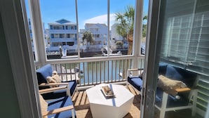 A glass-enclosed balcony with a white coffee table, blue cushioned seating, and a view of a beachfront property. - Canal home with access to the Intracoastal Waterway only steps from the beach! (Ocean Isle Beach)