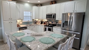 A kitchen with white cabinets, a stainless steel refrigerator, a microwave, and a granite countertop with green placemats. - Canal home with access to the Intracoastal Waterway only steps from the beach! (Ocean Isle Beach)