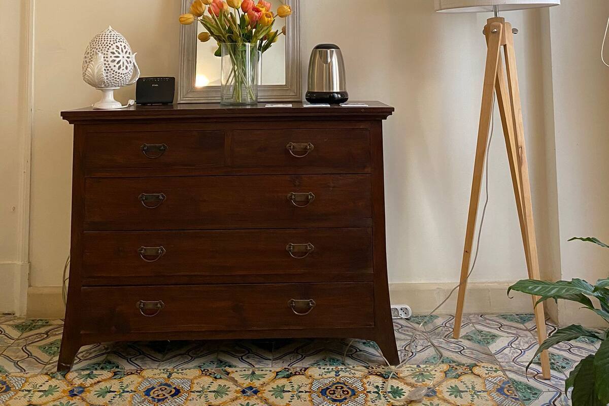 A room with a wooden dresser, a flat-screen TV mounted on the wall, a mirror, a vase with flowers, a lamp, and a tripod.