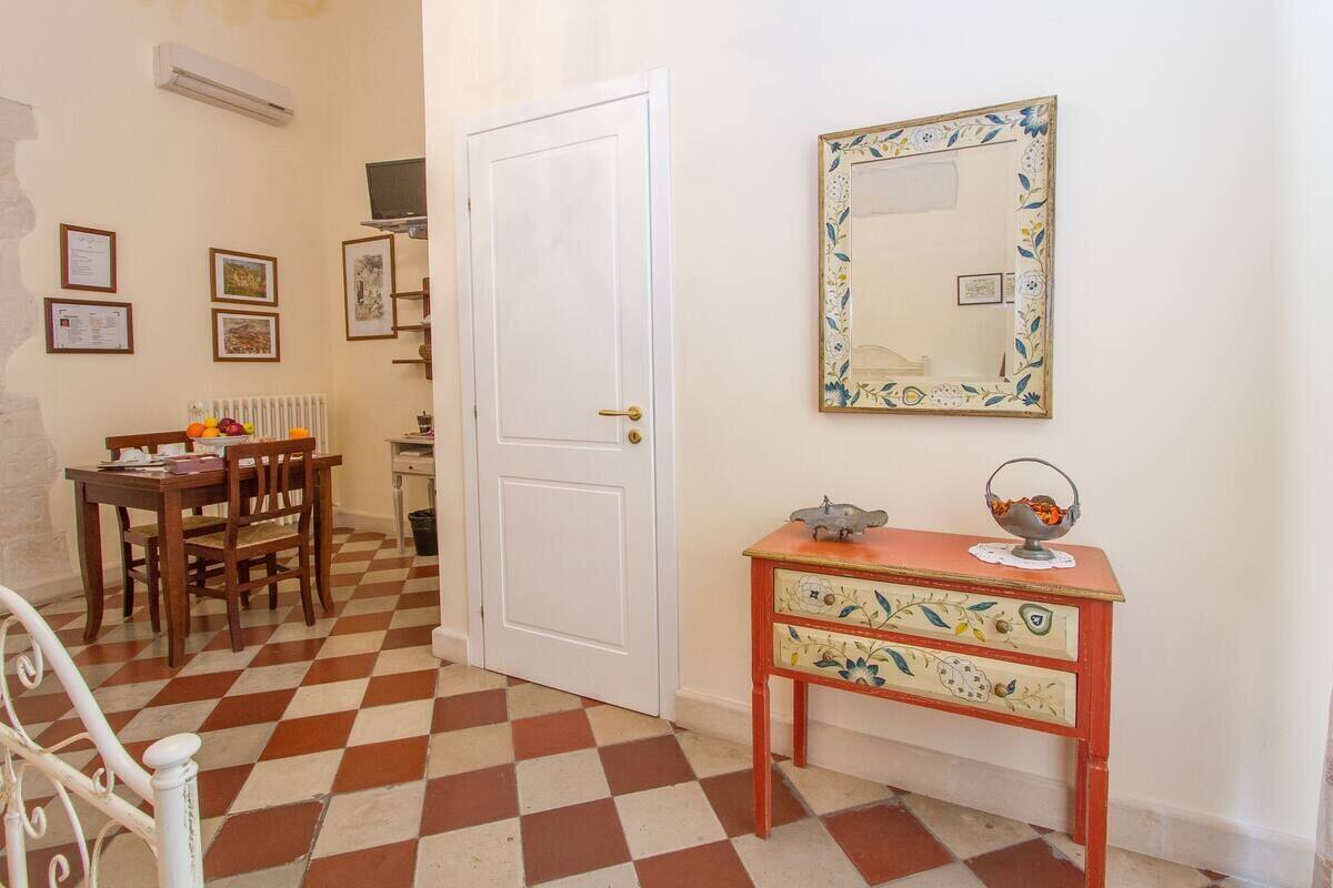 A room with a checkered floor, a white door, a wooden table with chairs, a small table with decorative items, and a wall-mounted mirror.