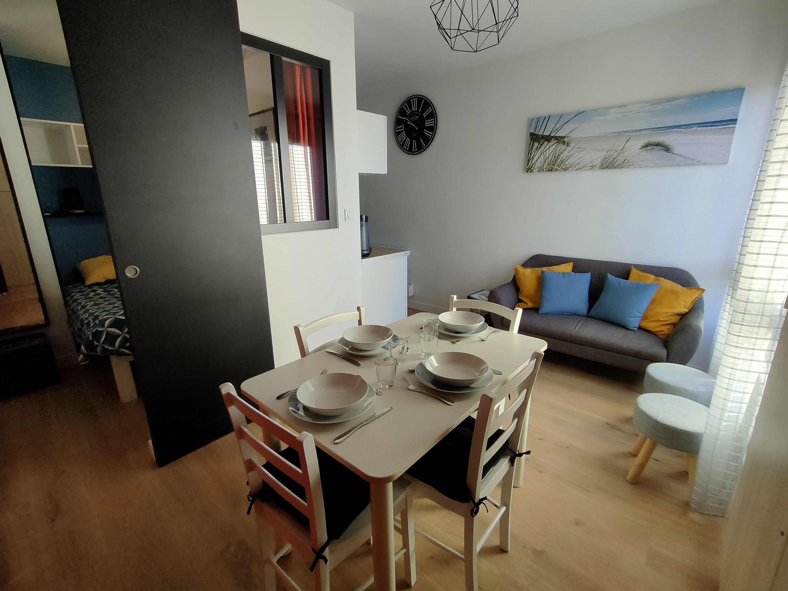 A dining area with a table set for four, a sofa with cushions, a clock, and a beach-themed painting on the wall.