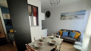 A dining area with a table set for four, a sofa with cushions, a clock, and a beach-themed painting on the wall. - Bel Appartement au Bord de L'océan (Saint-Jean-de-Monts)