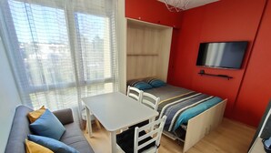 A compact room with a sofa, a small table, a bed with a TV above it, and a window with sheer curtains. - Bel Appartement au Bord de L'océan (Saint-Jean-de-Monts)