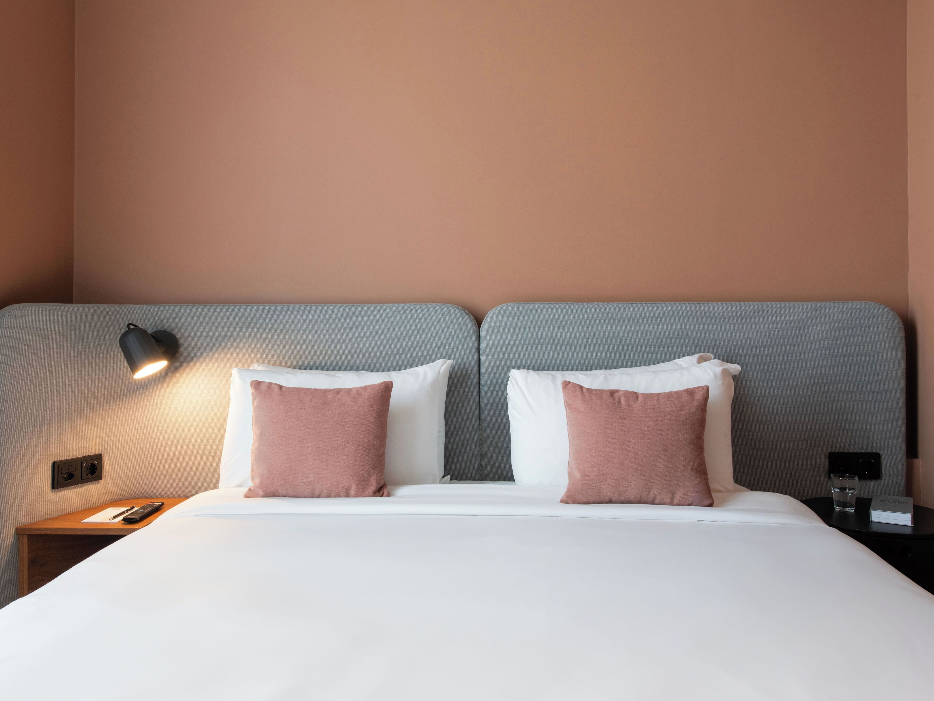 Habitación de hotel con cama, dos almohadas, mesita de noche, control remoto, vaso de agua y un libro.