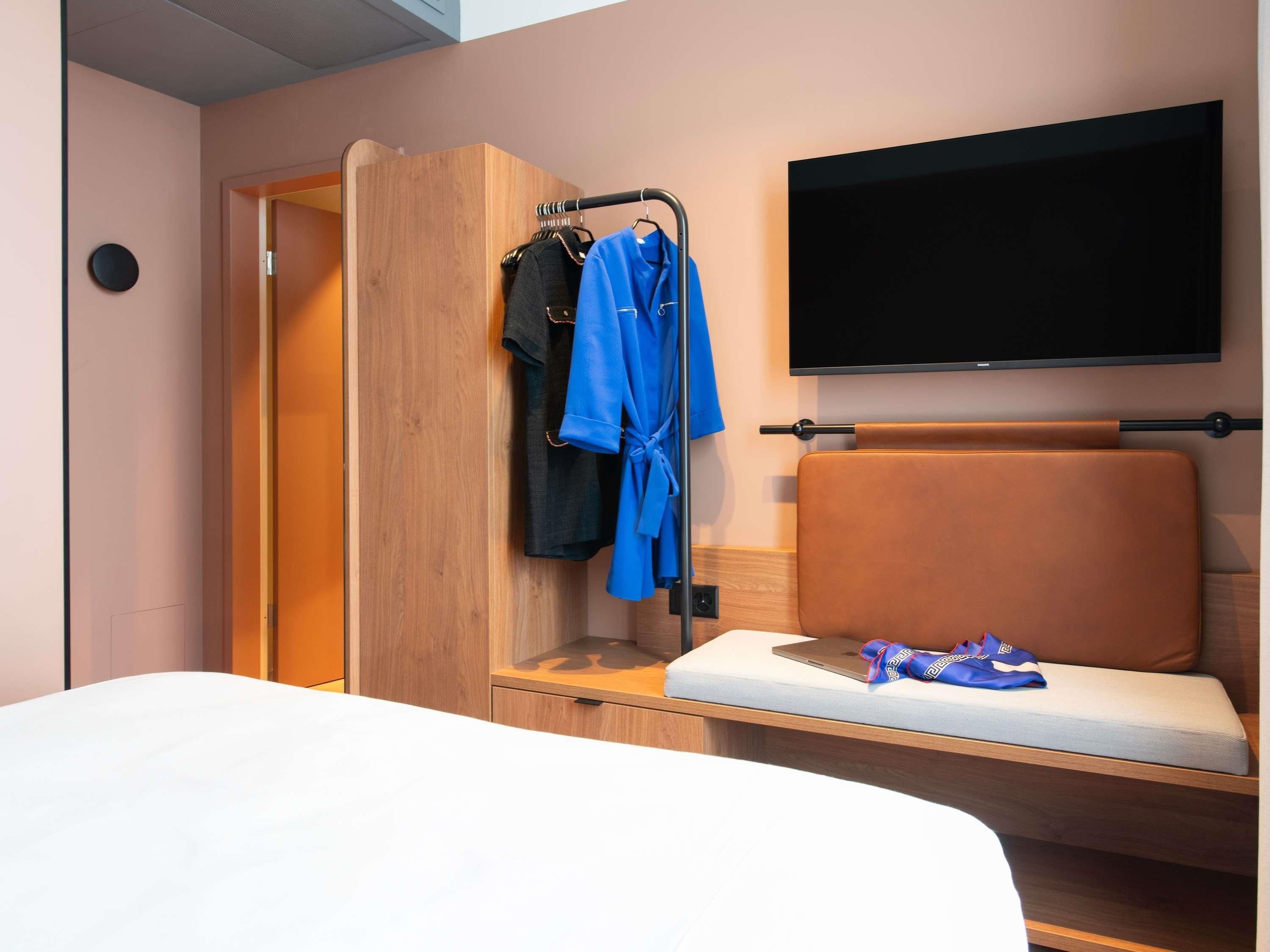 Une chambre d’hôtel comprenant un lit, un coin salon avec un coussin, un porte-manteau avec des vêtements, une télévision et une armoire en bois.