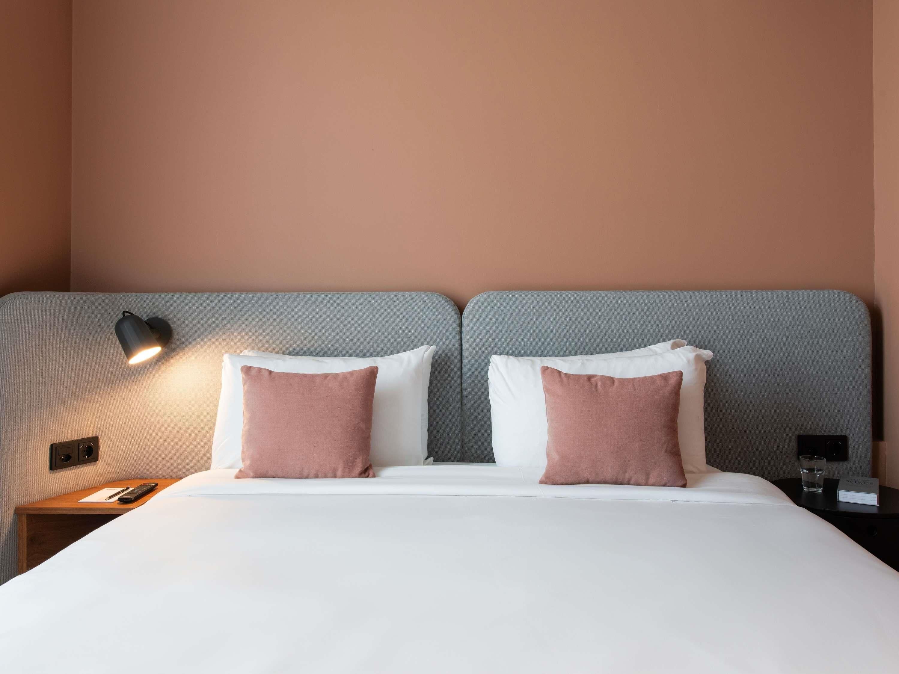 A double bed with two pillows, a bedside table with a remote control, and a wall-mounted lamp.