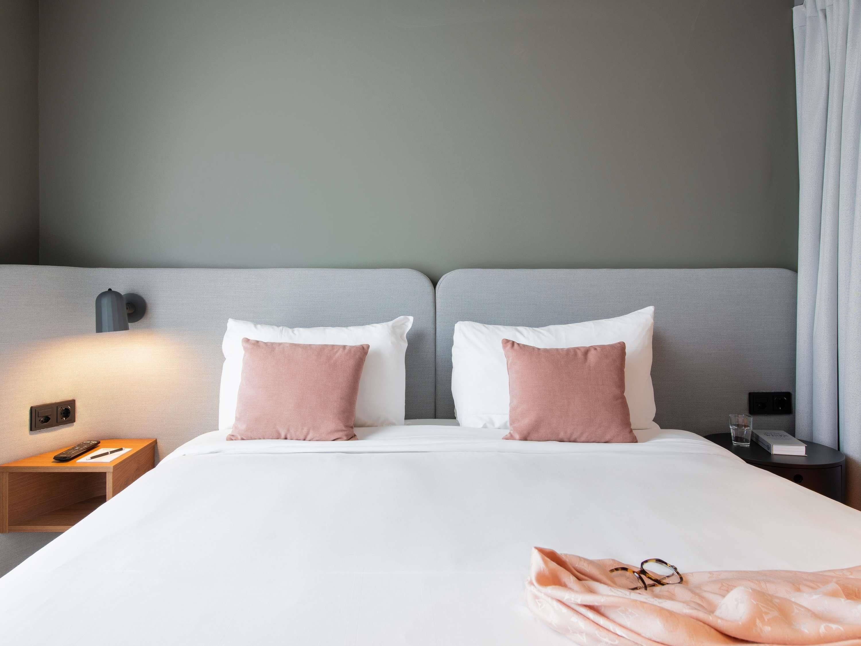 A hotel room with a bed, two pillows, a bedside table with a remote control, a nightstand, and a lamp.