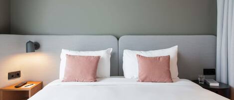 A hotel room with a bed, two pillows, a bedside table with a remote control, a nightstand, and a lamp.