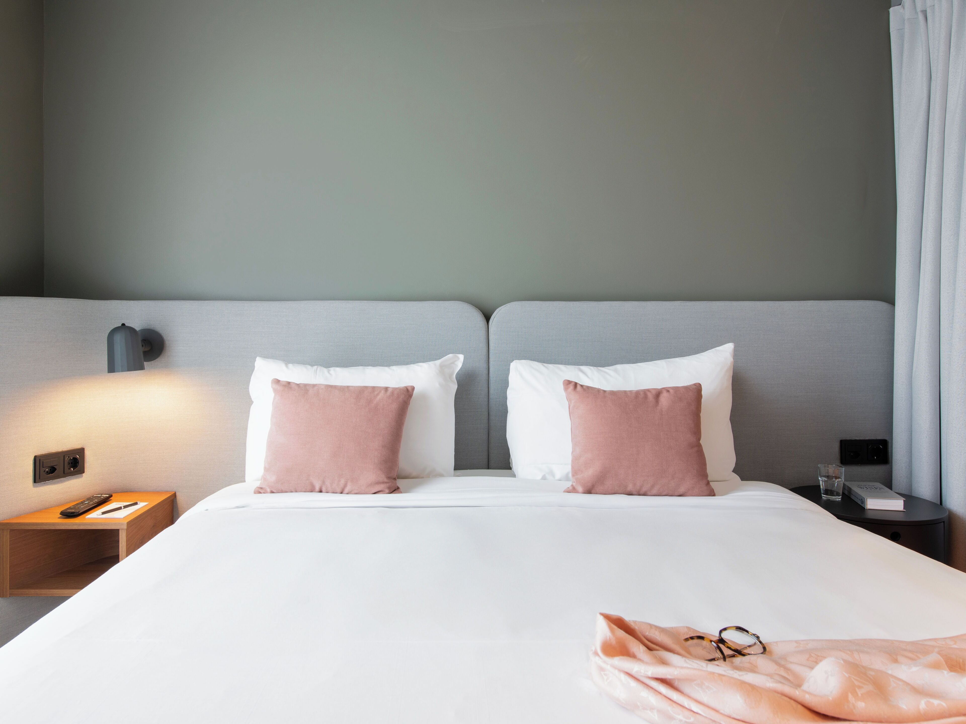 Hotelkamer met een bed, twee kussens, een nachtkastje met een afstandsbediening, een glas water en een boek.