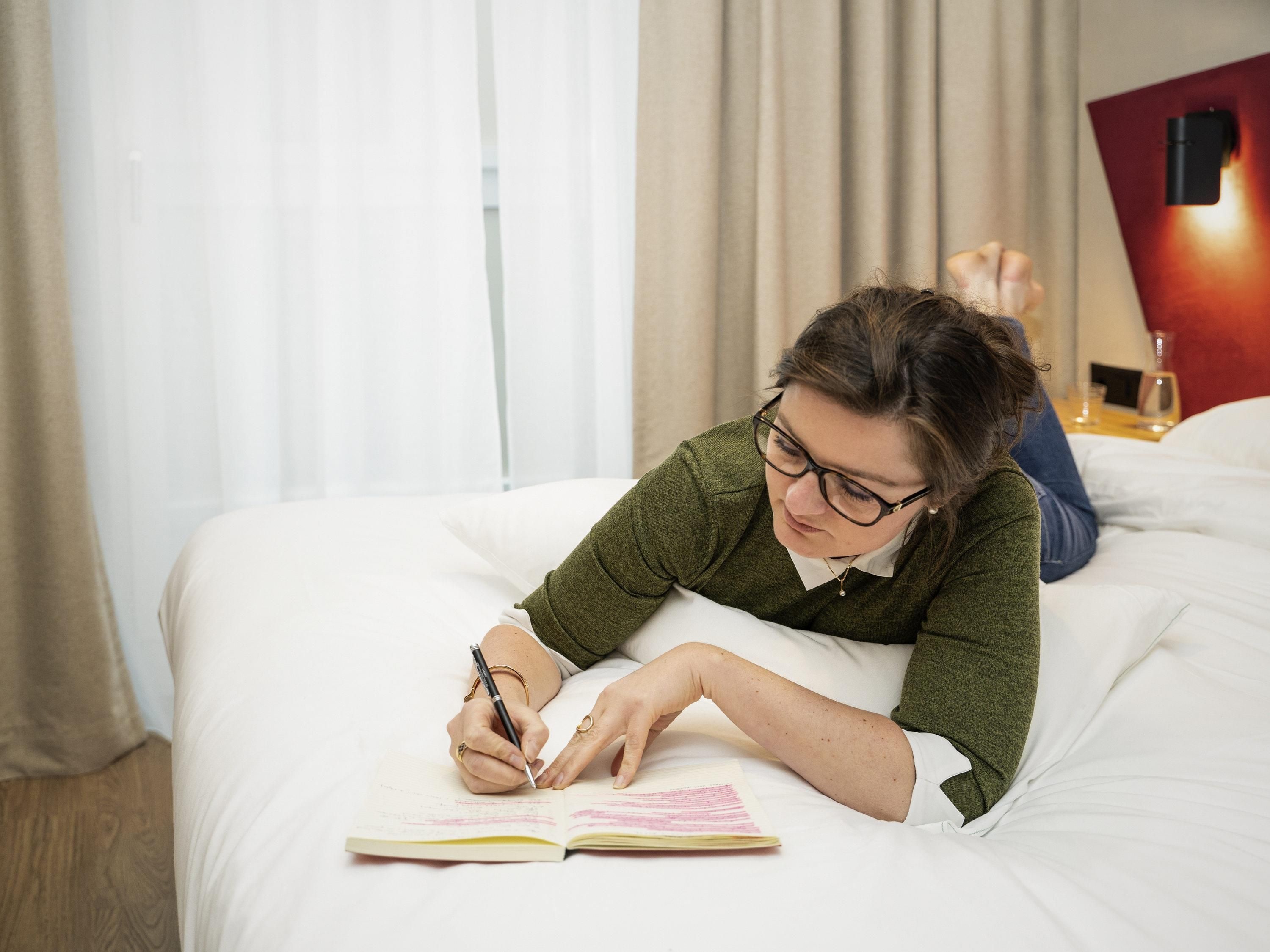 A person lying on a bed with a book, writing in a notebook.