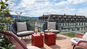 A terrace with wicker furniture, a red table, and a view of buildings and mountains. - Aparthotel Adagio Zurich Center (Zürich)