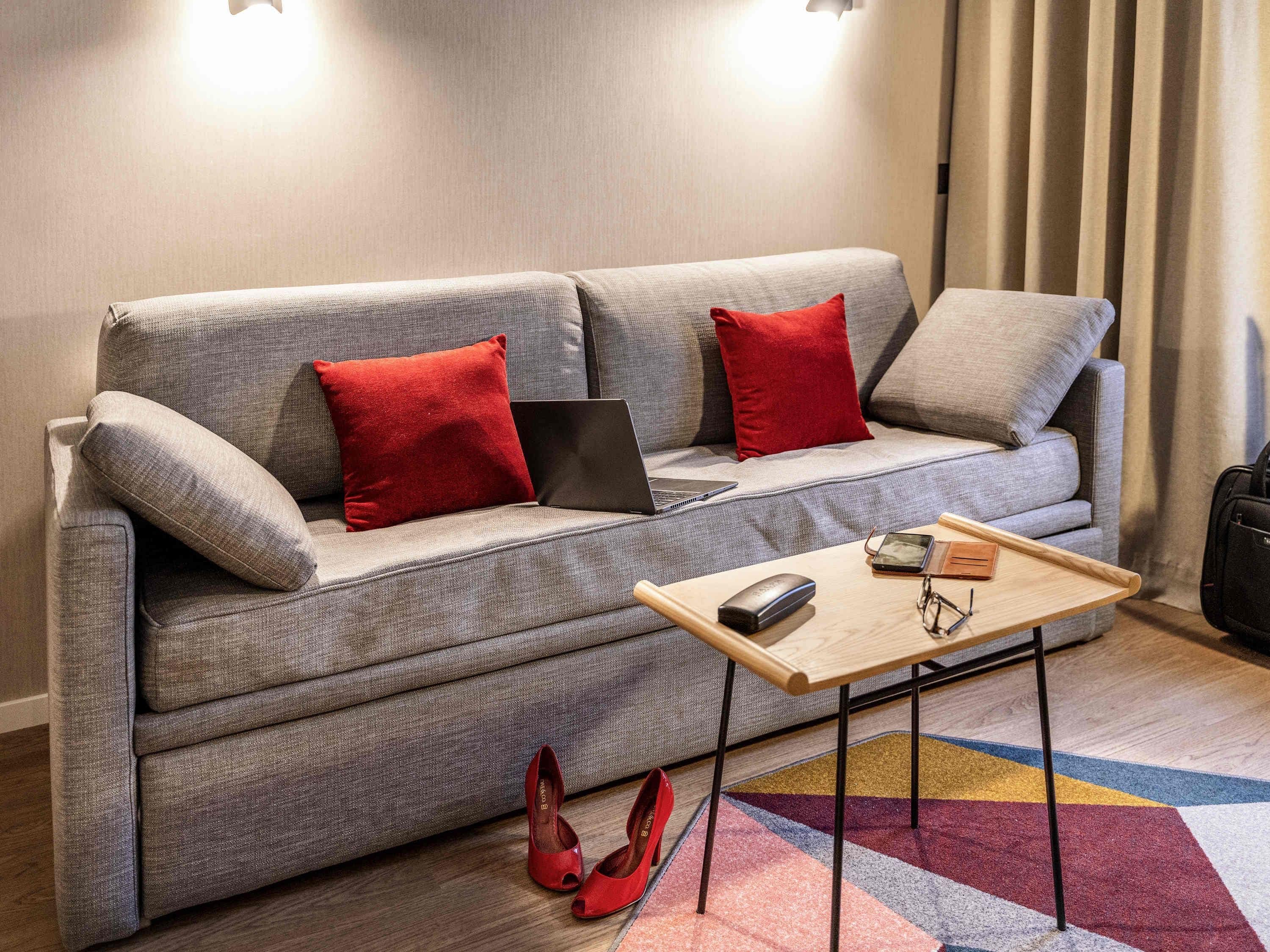 A modern living room with a grey sofa, red cushions, a laptop, a wooden coffee table, and red high-heeled shoes on the floor.