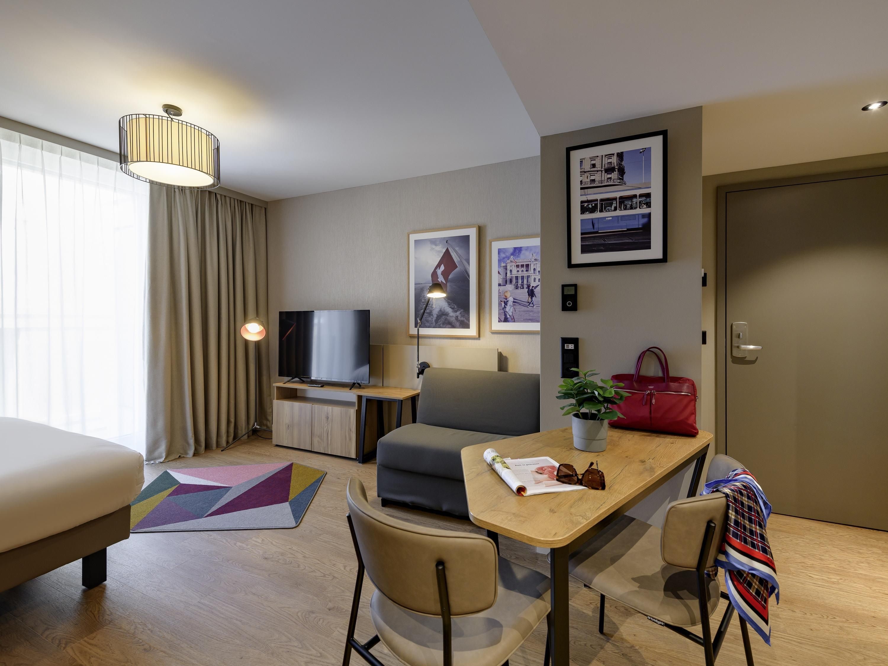 Habitación de hotel moderna con sofá, una mesa de comedor con sillas, un televisor, una planta en maceta y un bolso rojo.