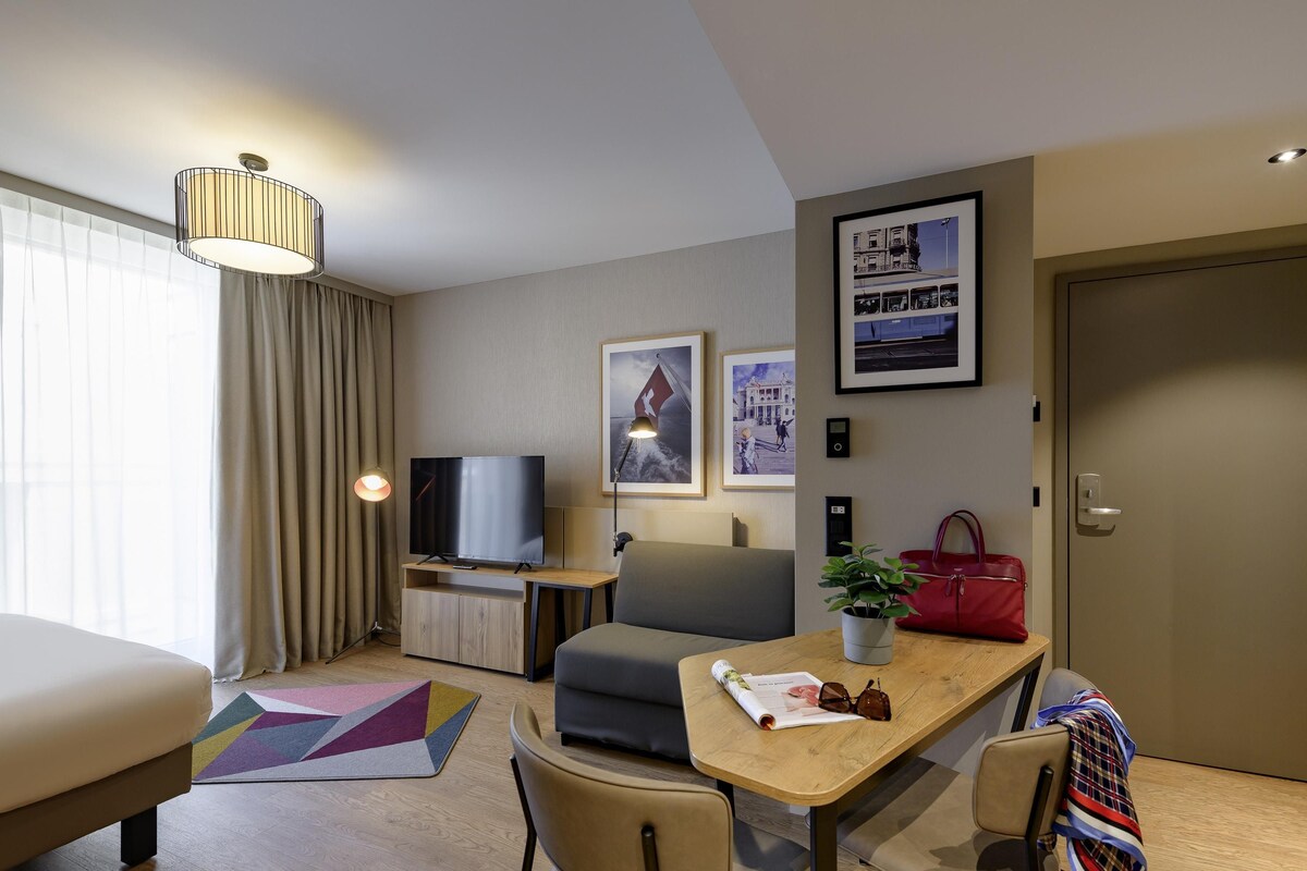 A modern hotel room with a sofa, a dining table with chairs, a television, a potted plant, and a red handbag.