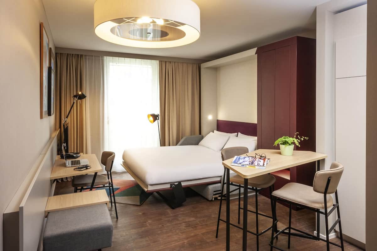 Habitación de hotel con cama, escritorio, sillas, lámpara y planta en maceta.