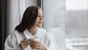 A person in a white robe holding a cup, sitting by a window with a view of a building. - Aparthotel Adagio Zurich Center (Zürich)