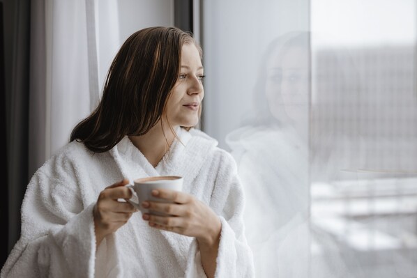 A person in a white robe holding a cup, sitting by a window with a view of a building. - Aparthotel Adagio Zurich Center (Zürich)