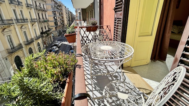 Un balcon avec une table et des chaises en métal, des plantes en pot et une vue sur une rue bordée d’immeubles.
