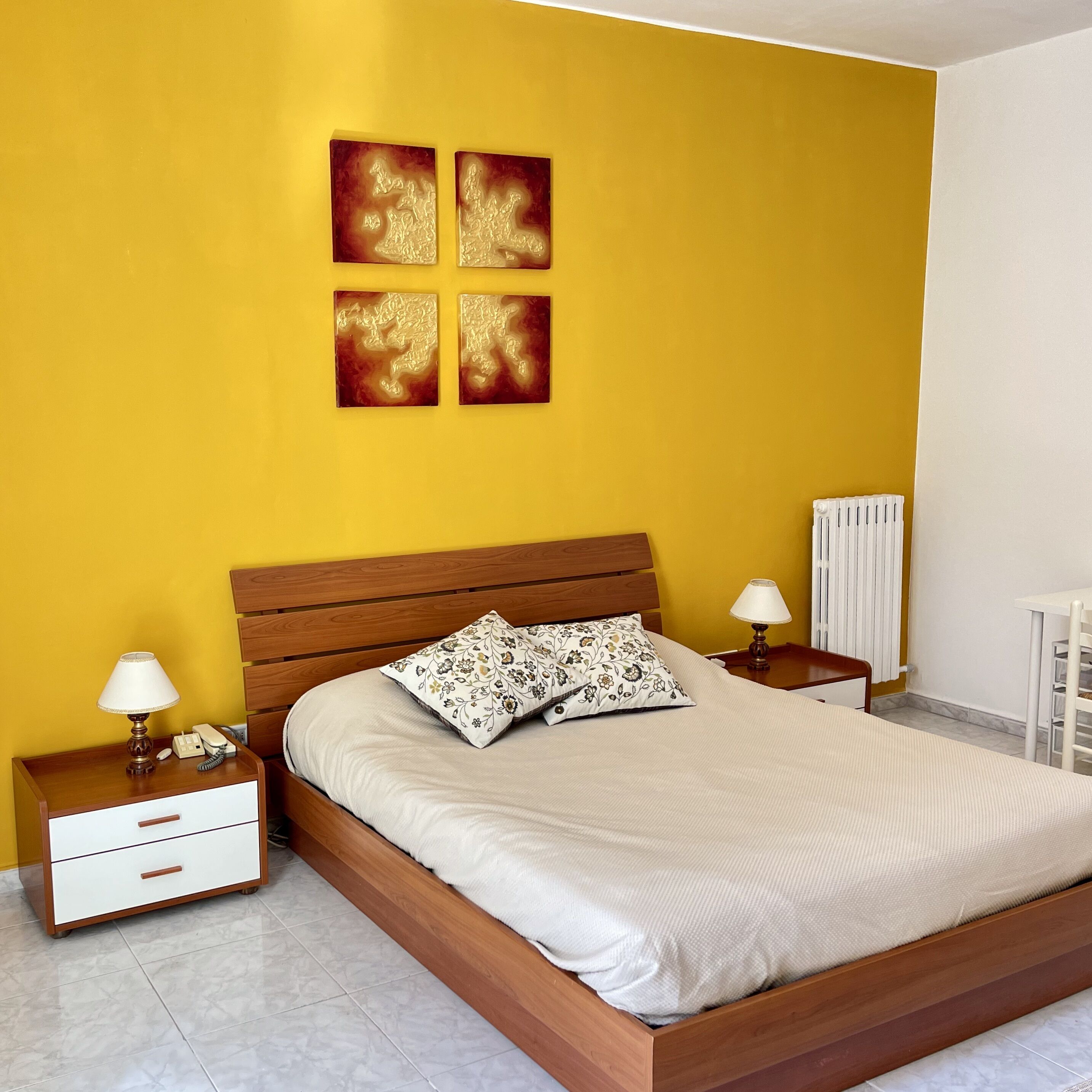 Un dormitorio con una cama de madera, dos mesitas de noche, una mesita blanca, un radiador y una pared amarilla con cuatro obras de arte.
