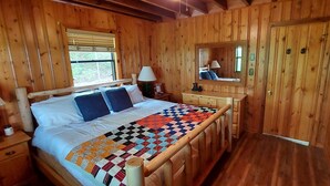 Un dormitorio de cabaña de madera con cama, mesita de noche, cómoda, espejo y ventana.