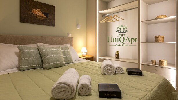 Ein Hotelzimmer mit Bett, Kissen, einem Gemälde, einem Nachttisch, einer Lampe, einem Regal mit Körben und dem Logo für UniQapt Confu Greece.