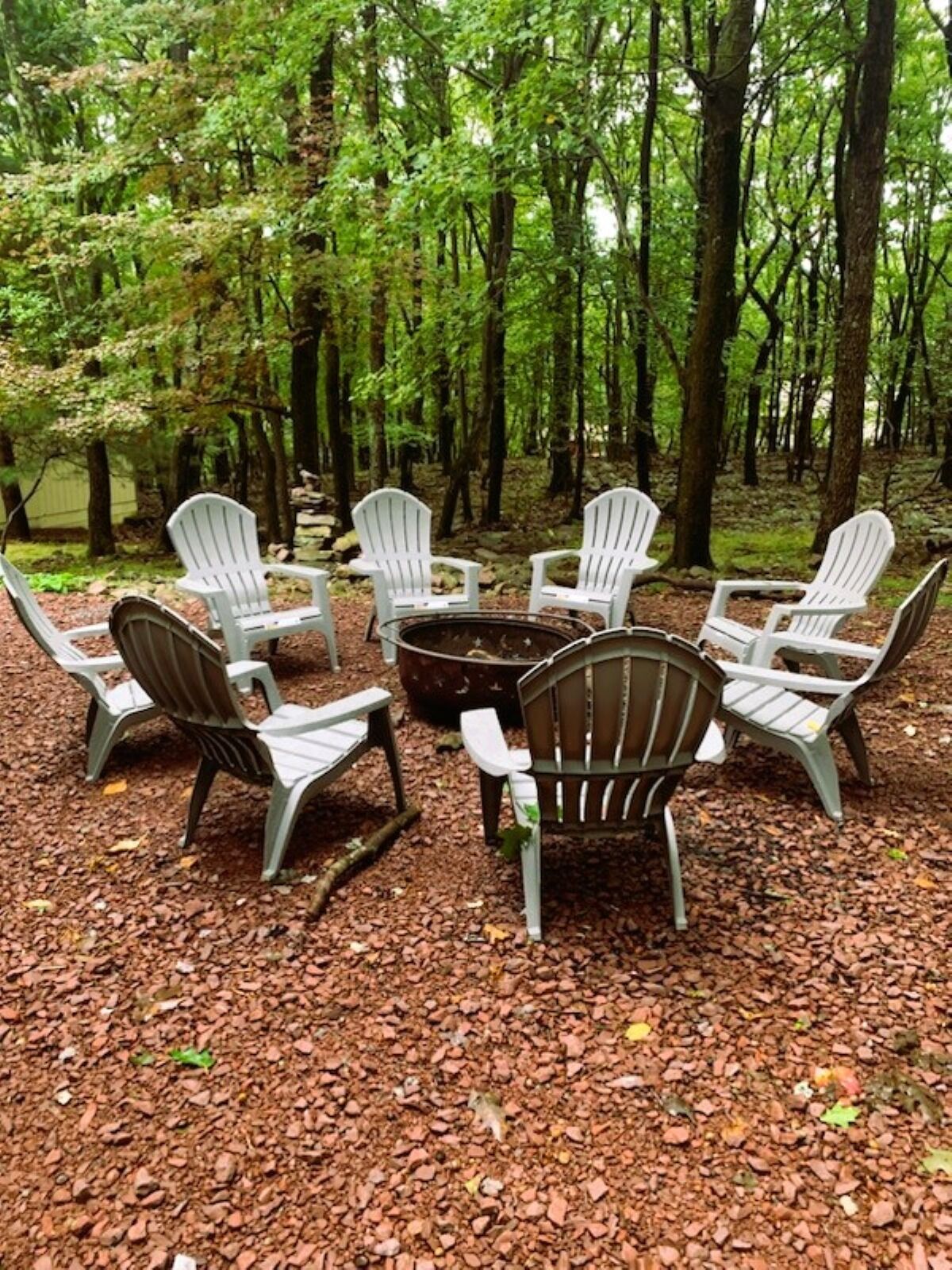 Un aménagement circulaire avec des chaises Adirondack disposées autour d’un foyer central, le tout situé dans une zone boisée.
