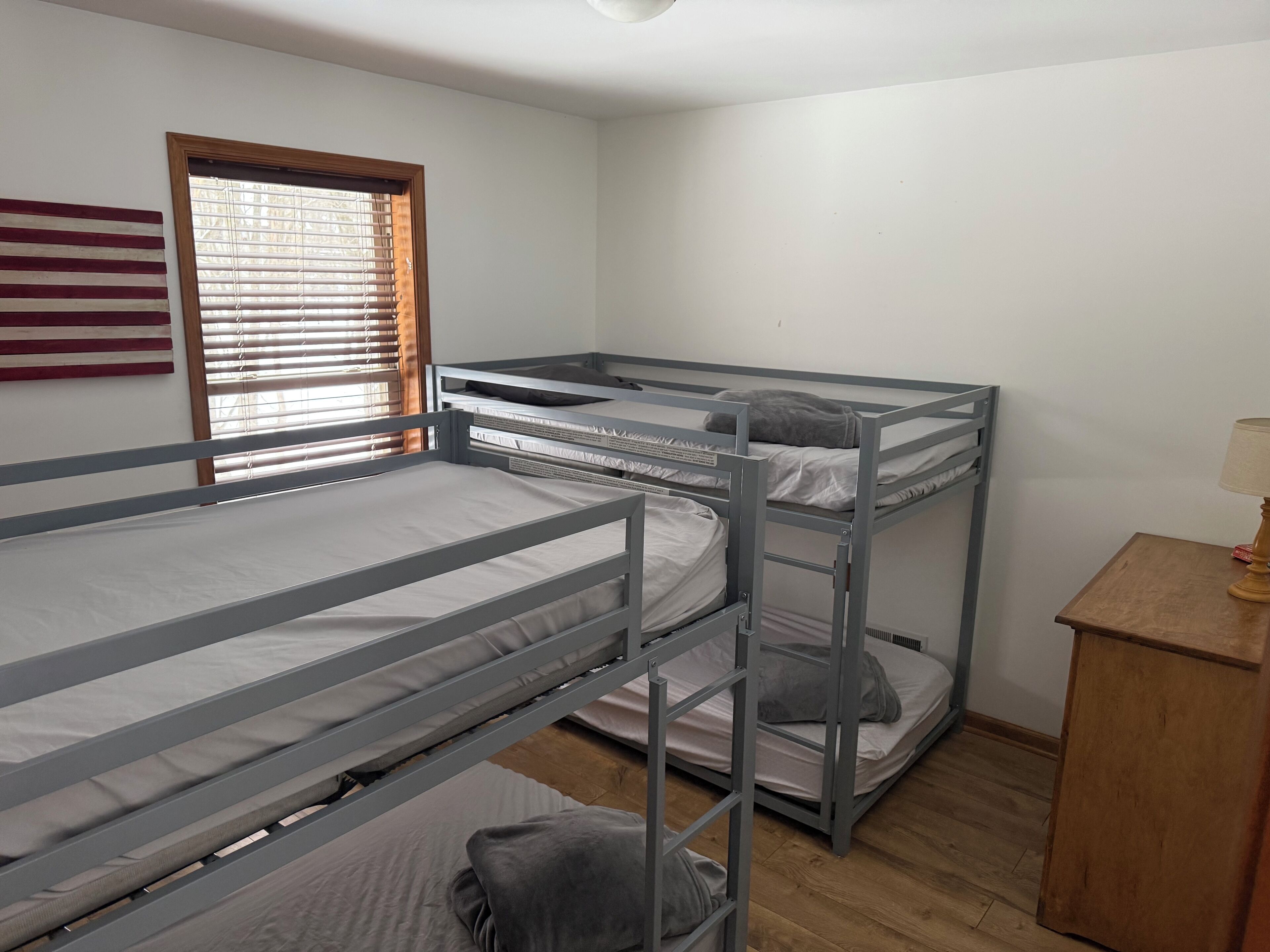 A bunk bed room with a wooden floor, a window with blinds, and a small wooden table with a lamp.