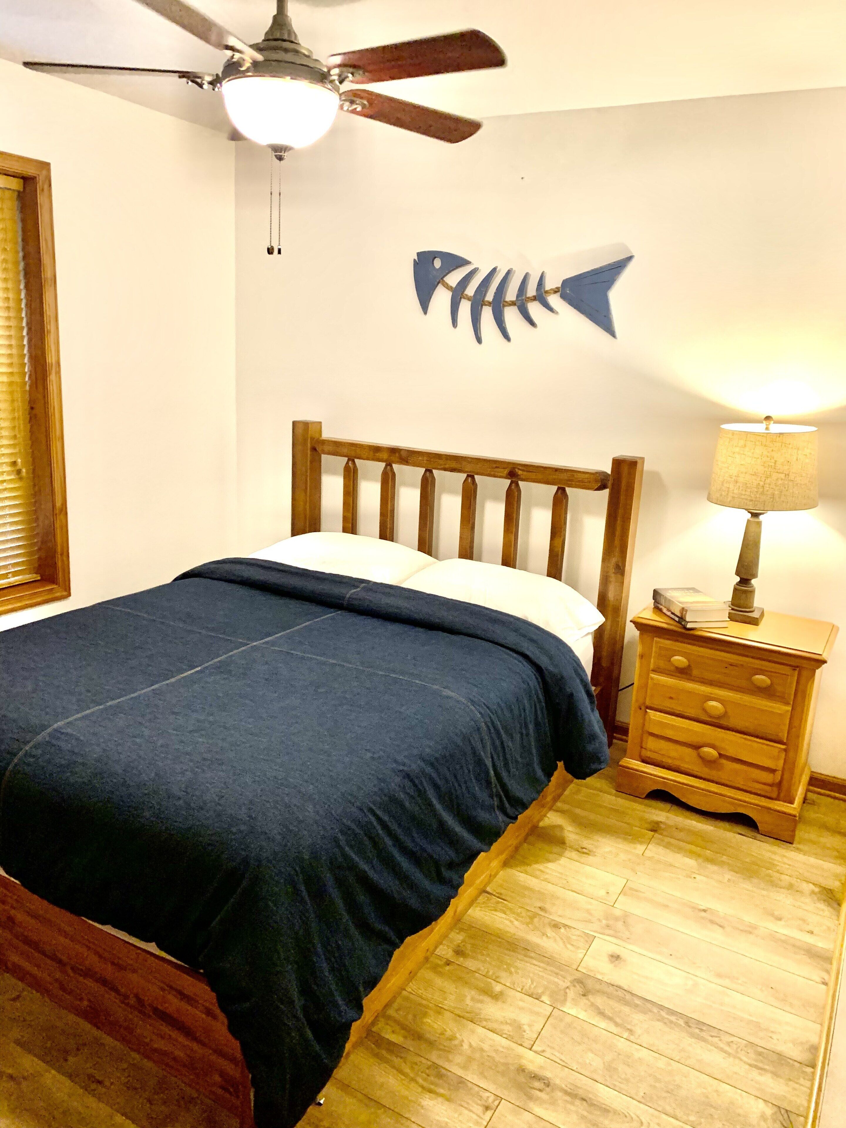 A bedroom with a wooden bed, a nightstand with a lamp, a ceiling fan, and a wall decoration of a fish.