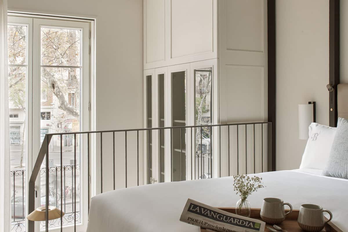 A hotel room with a large bed, a wooden tray with a newspaper, a vase with flowers, and a view of the street through the window.