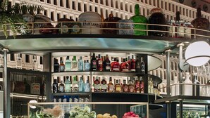 Área de bar com exposição de garrafas e vitrine com frutas e legumes. Há duas cadeiras de vime na frente.