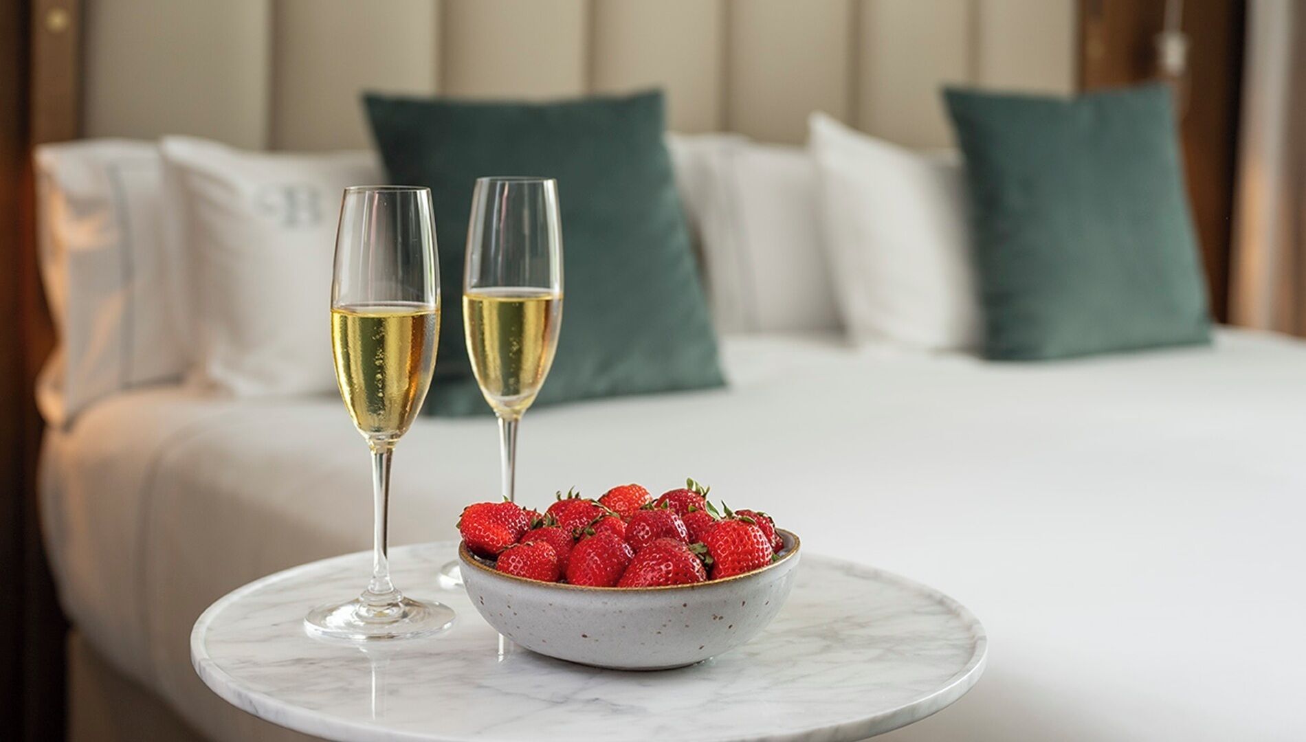 A bed with white bedding and green pillows, a bowl of strawberries, and two glasses of champagne on a marble table.