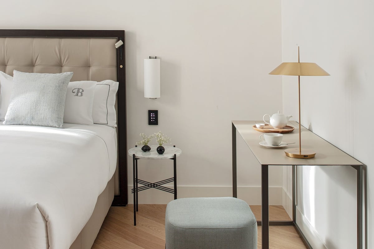 A modern hotel room with a large bed, a bedside table with a lamp, a small side table with decorative items, and a window with a view.