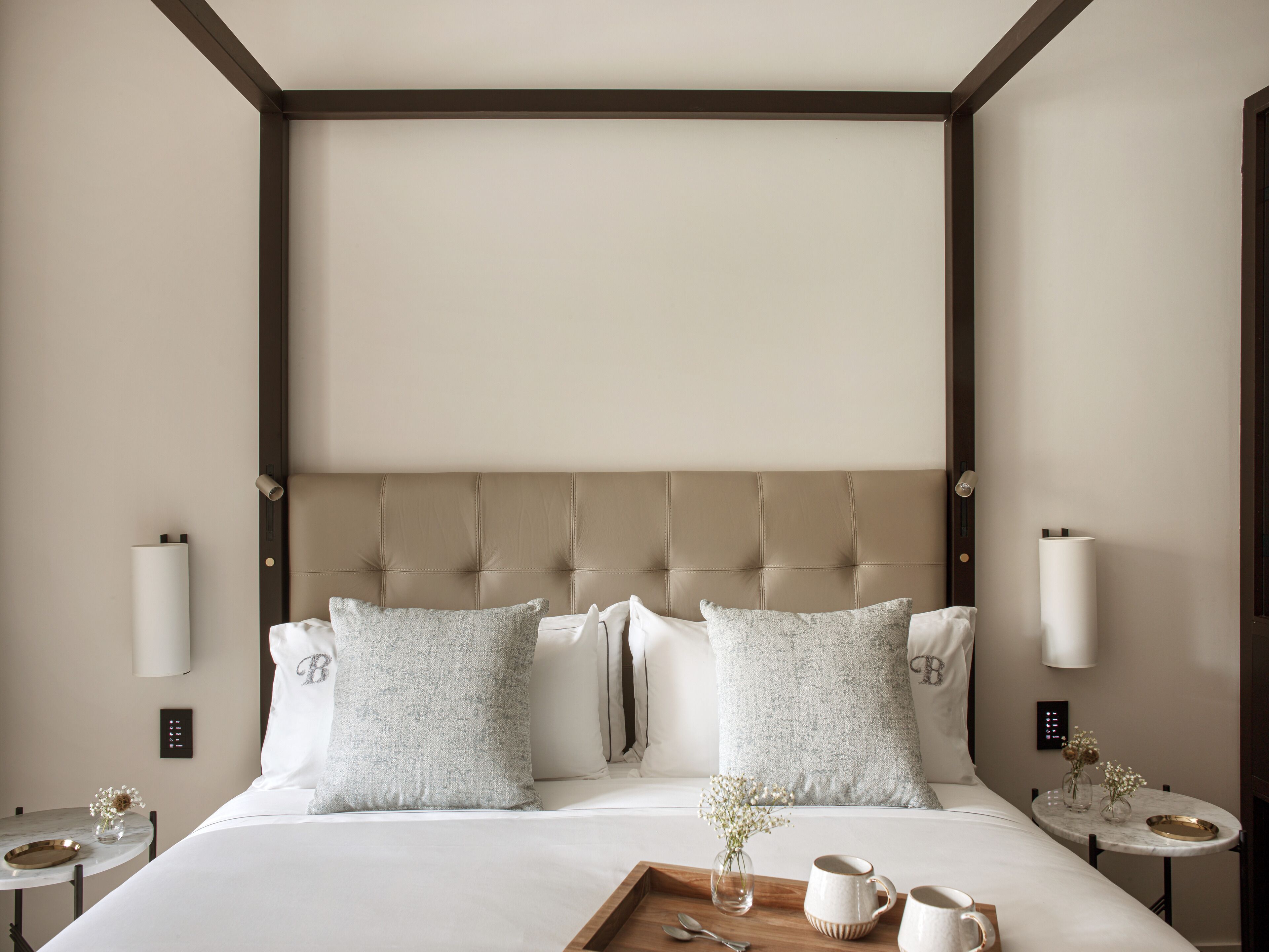 Een hotelkamer met een gestoffeerd hoofdeinde, een houten nachtkastje met dienblad, een vaas met bloemen en een bedlamp.