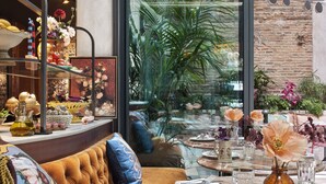 Uma área de jantar acolhedora com uma mesa redonda, um banco estofado e uma grande janela com vista para uma parede de tijolos e vegetação lá fora.