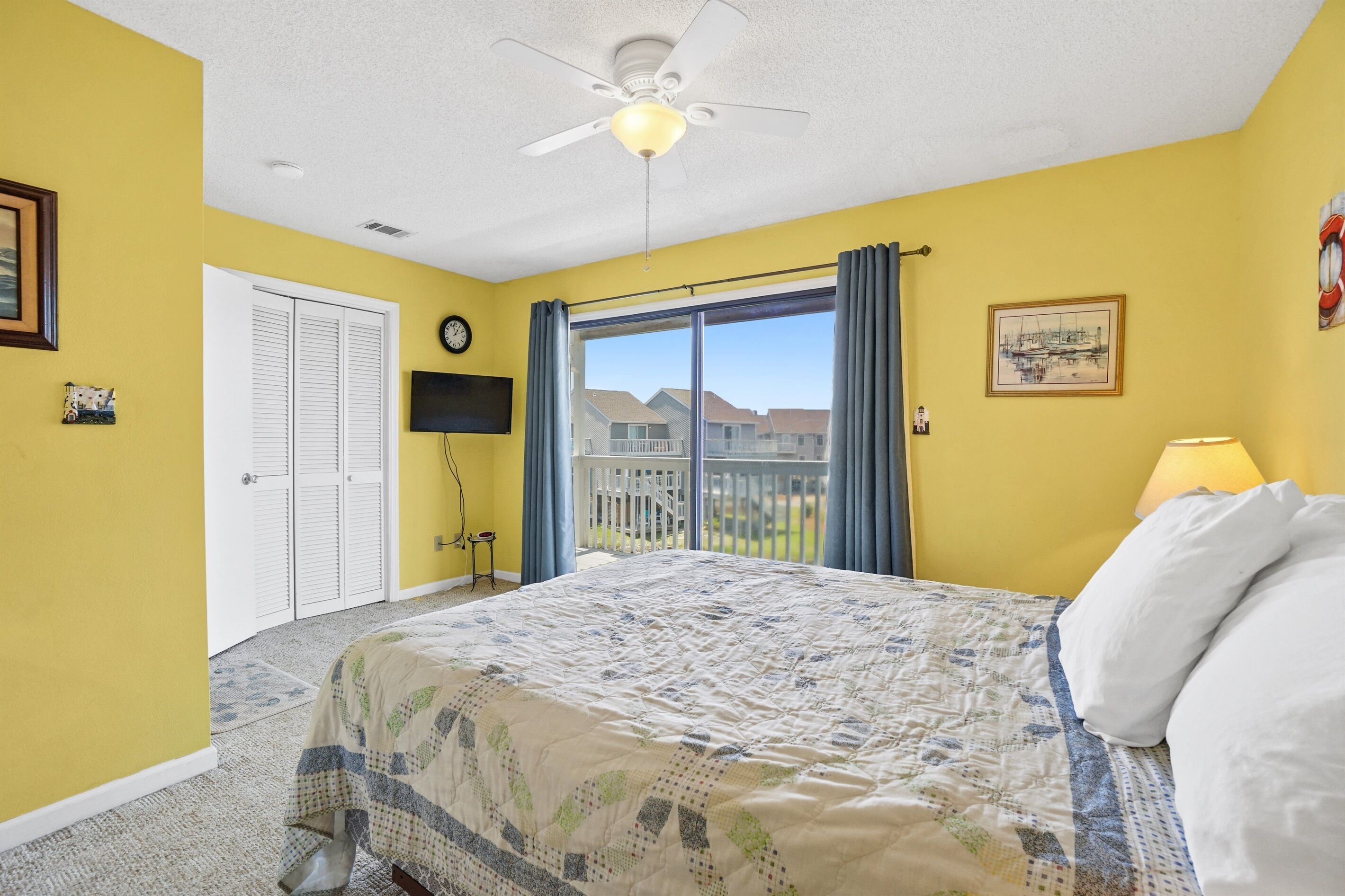 A bedroom with a large bed, a ceiling fan, a window with curtains, a door, and a wall clock.