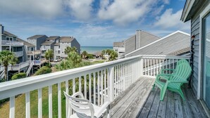 Una terraza de madera con barandas blancas y dos sillas Adirondack, con vista a una zona residencial con casas y una playa.