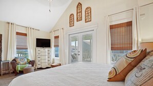 A bedroom with a large bed, a television, a dresser, and a chair. There are three windows with curtains, and a balcony door. - Fall 2026 Rate Drop! 3 BR/3 BA Cottage -Dog Friendly! (Port St. Joe)