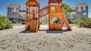 Eine hölzerne Spielanlage an einem Sandstrand, im Hintergrund Wohnhäuser.