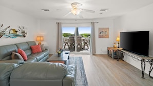 Un salon comprenant un canapé, une table basse, un téléviseur et un ventilateur de plafond. Une porte-fenêtre coulissante donne accès à un espace extérieur aménagé avec une table et des chaises.