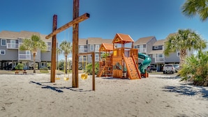 Un terrain de jeux au bord de la plage, avec un toboggan et des balançoires, entouré d’immeubles à plusieurs étages dotés de balcons.