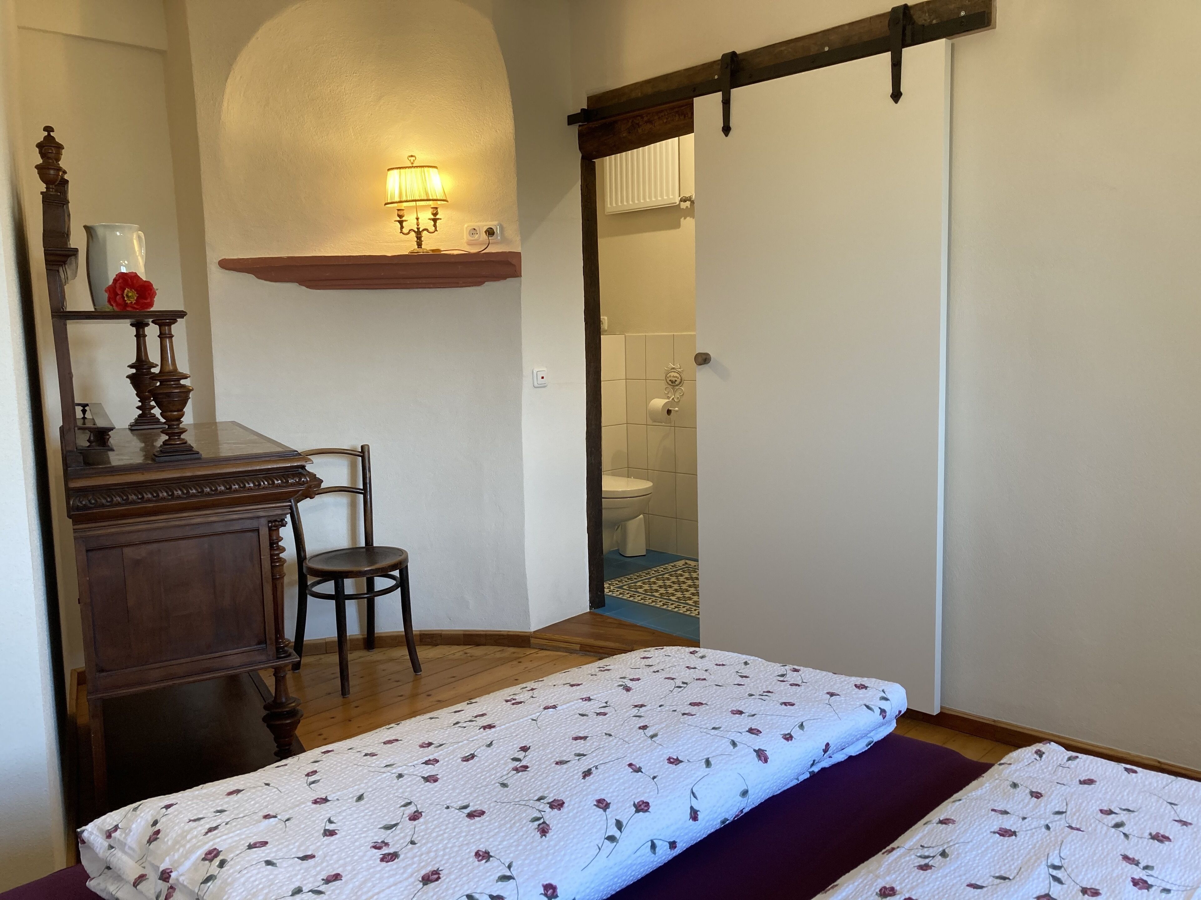 Un dormitorio con cama, una silla, un escritorio con lámpara y un baño visible a través de una puerta abierta.