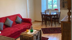 A living room with a red sofa, a wooden coffee table, and a chandelier. There is a dining area with a table and chairs, and a window with a view of greenery. - Vacation at the winery (Maring-Noviand)