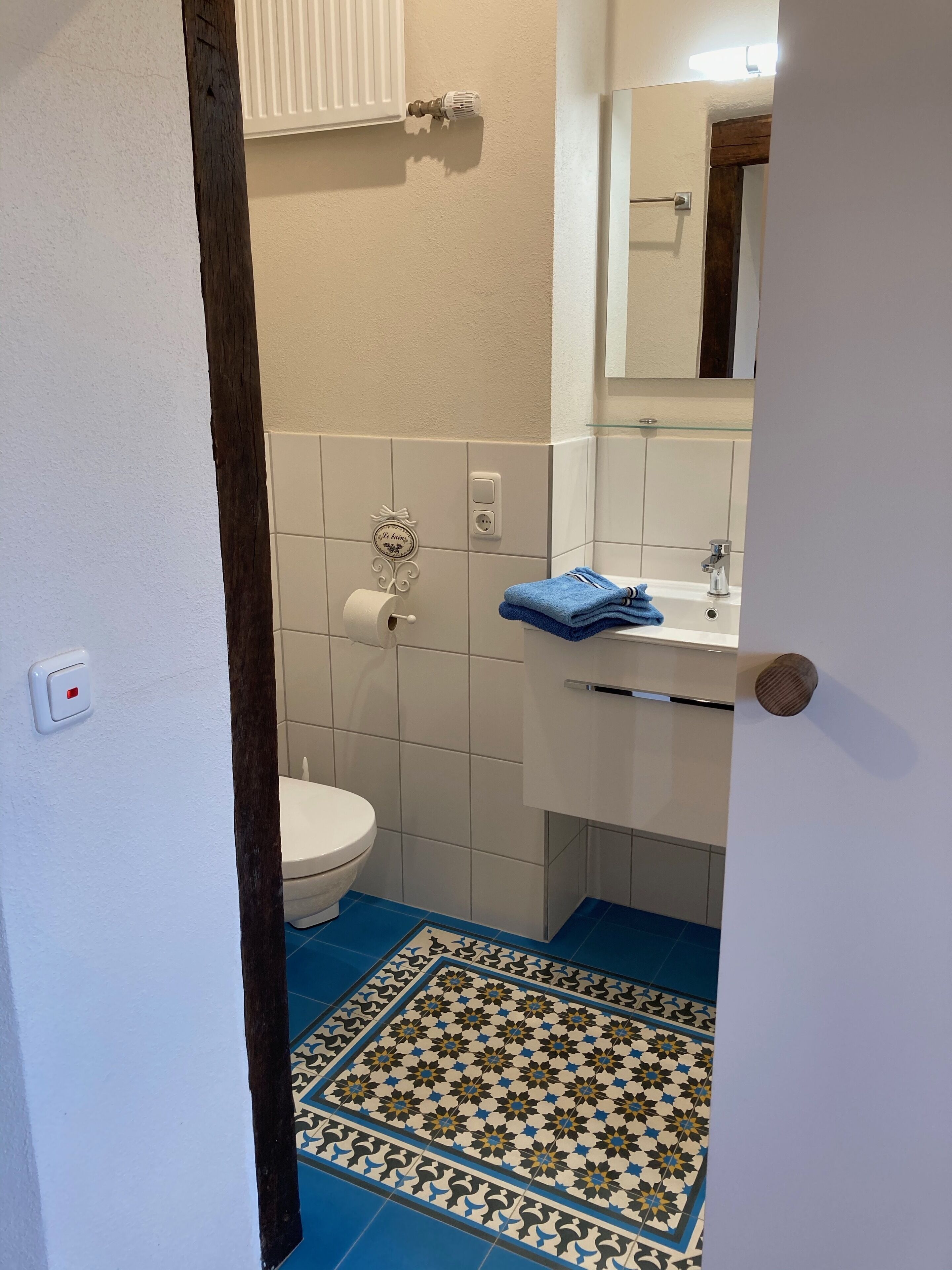 Un baño con inodoro, lavamanos y espejo. Hay un felpudo azul con estampado y una toalla blanca sobre el lavamanos.