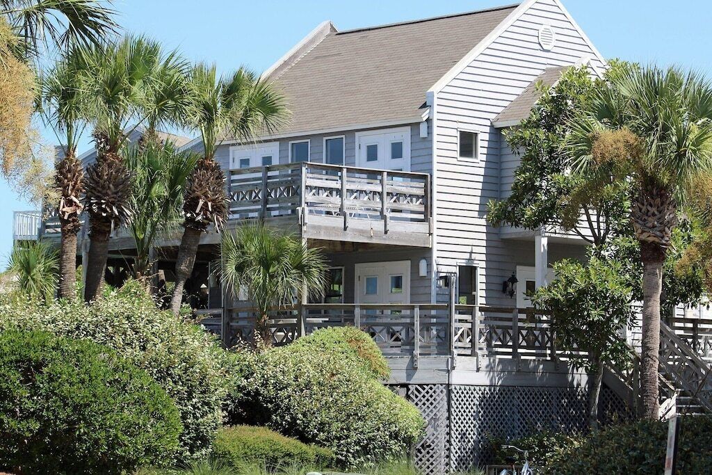 A two-story building with a balcony, surrounded by palm trees and shrubs.