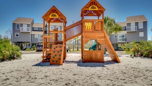 Una estructura de juegos de madera en una playa arenosa, con edificios residenciales altos de fondo.