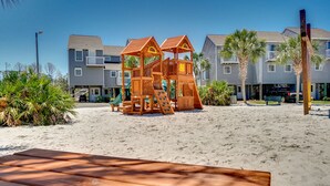 Una estructura de juego de madera en una playa arenosa, con un banco en primer plano y un edificio de varios pisos al fondo.